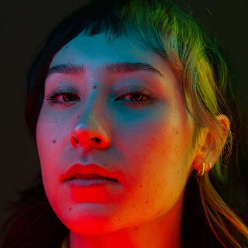 A closeup of kira shiina in which their face is lit with red and blue and yellow and purple