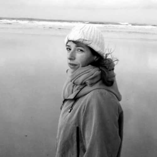 Isabelle Launay wearing a windbreaker and a knit hat standing in front of a still body of water in black and white
