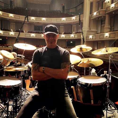 Marc Dicciani sitting onstage on the edge of a drum set with theater tiers and boxes behind the set