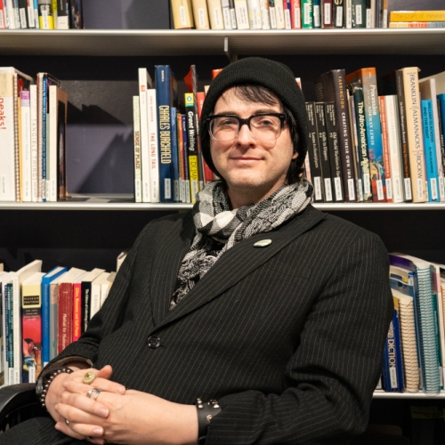 headshot of marshall oneill against a background of books. marshall is wearing a black pinstripe blazer, a black and white patterned scarf, a black beanie, and a studded leather bracelet. marshall has a soft smile and is wearing black glasses. 