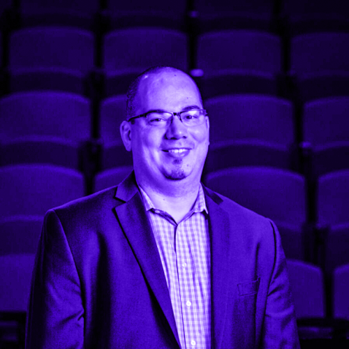 portrait of micah jones overlaid with violet. micah is sitting in auditorium risers wearing a blazer and a checkered collared shirt smiling with a little goatee and semi-rimmed glasses.