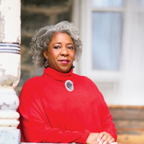 Monna Morton stands on a porch with her hands folded in a red turtle neck sweater, earrings and a brooch.