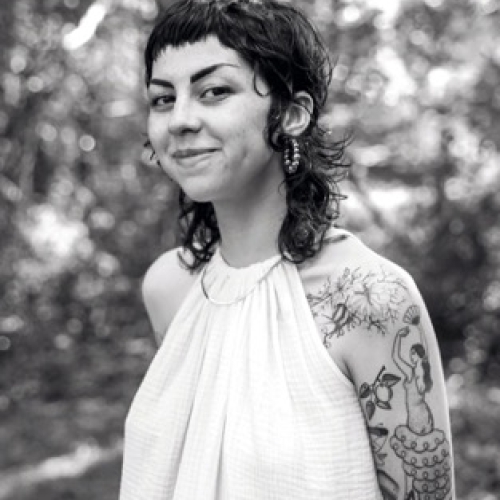 black and white portrait of mia Elise Paltrow-Murray with a curly banged black mullet and a white top. mia's left arm is covered in tattoos. mia has a sly smile and sharp eyebrows 