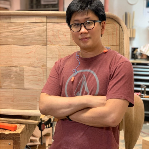 Keunho Peter Park in a rose-colored T-shirt, standing with arms folded in front of a piece of unfinished furniture.