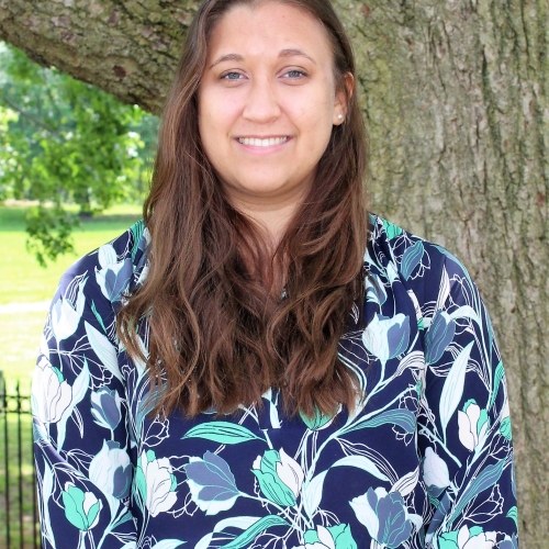 Rachel Kiskaddon smiling and standing in front of a tree and green background. She is wearing a floral shirt with dark blue, teal, and white streaks.