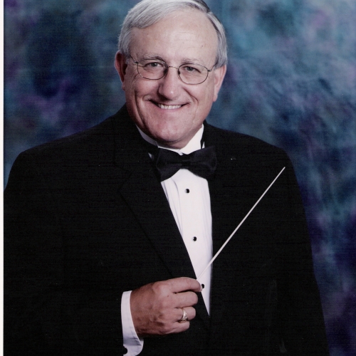 Photo portrait of john lindner smiling in a tuxedo against a blue watercolor-like photo backdrop, holding a conductor's rod. 