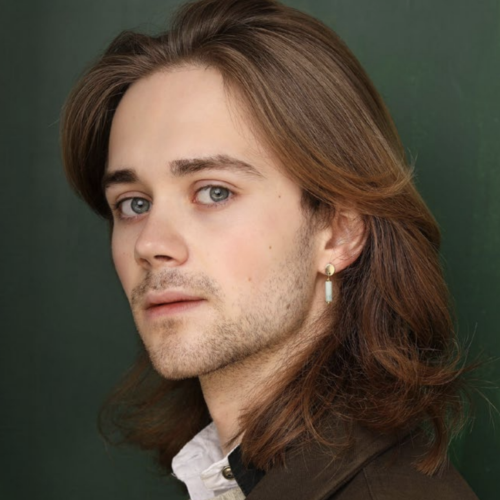headshot of jack hopewell in partial profile, glancing at the viewer with soft green eyes. jack has long flowing hair and a tamed scruff on his face. the background is a green matching jack's eyes