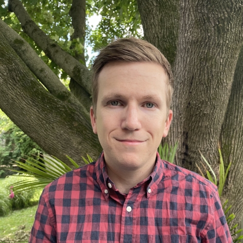 headshot of Timothy Potts, pictured wearing a black and red checkered collared shirt against a background of a tree in a lush garden. timothy is smiling, and has dark blond hair swept to the side. 