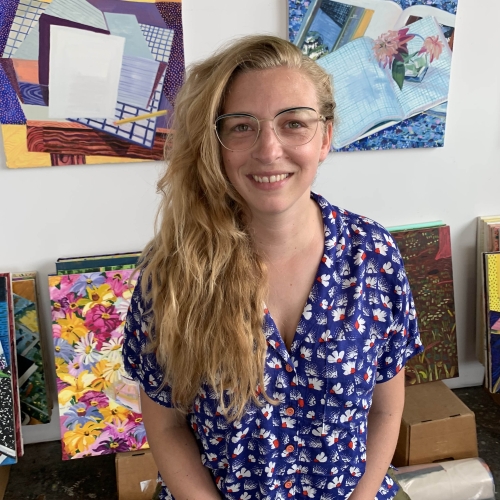 Lauren Whearty wearing a bright blue shirt with a print of white flowers on it and standing in front of a number of her colorful paintings that are both on the wall and stacked behind her