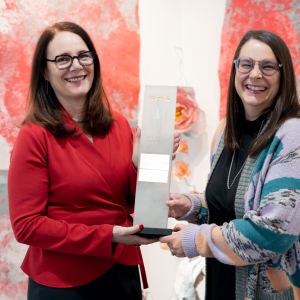 sheryl oring and jessica kahle stand side by side looking at thie viewer holding a trophy against a background of splatter-pattern artworks. 