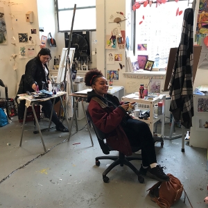 Two students work on their art in the painting studios. One student smiles at the camera while they work.