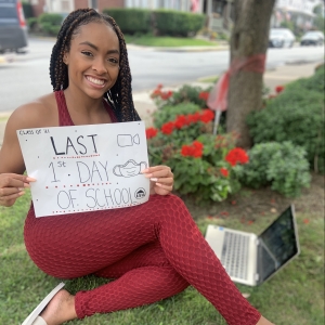 Photo of Azari sitting on the grass with their laptop, holding a sign that reads "Last First Day of School"