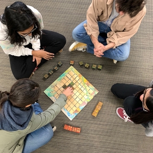 Students sit around a game board and play a game with small square wooden pieces.