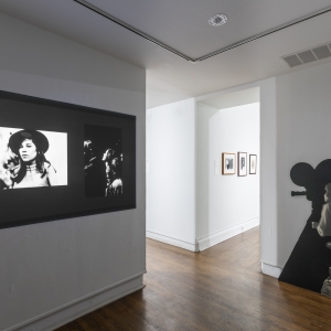 Invisible City Exhibition, Gallery D: film projection onto a dividing wall showing two still frames of people to the left and a black and white cut-out of a woman standing with a camera 