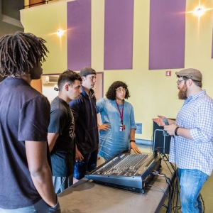 Students discuss audio production around a mixing board