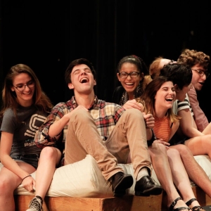 Theater students sit on a bed on stage and laugh during a performance