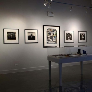 Installation view of Joel-peter Witkin: From The Studio at Catherine Edelman Gallery. Several photographs are pictured hanging on the white, gallery walls; a table in the foreground has objects under a plexiglass case.