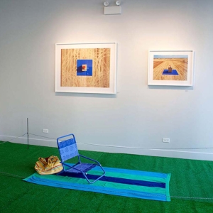 Installation view of What is Home? At Catherine Edelman Gallery. Photographs adorn the white gallery walls in the background; a green, artificial turf carpet with a multicolored beach towel and blue beach chair are set up on the floor, surrounded by a thin cable. A sign reading “What is Home?” is on the far left wall.