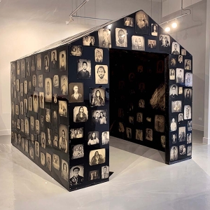Keliy Anderson-Staley’s “Shelter in Place,” a large constructed house-like building made from blackened aluminum sheathing is covered in 560 tintypes, looms against white gallery walls in this installation view of “What is Home?” at Catherine Edelman Gallery.