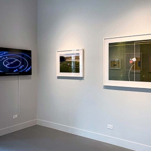 Installation view of What is Home? At Catherine Edelman Gallery. Two framed and mounted photographs adorn the white gallery walls on the right, while a black screen displays a still image from a multimedia work.