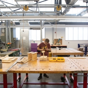 Interior of UArts makerspace classroom. a person with glasses is assembling a wooden object on a large worn wooden table. the room if lofty and bright. 