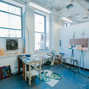 Interior of a painting studio, with various small paintings on easels, tables, and leaned against the wall. a partially painted canvas is on the floor in the middle of the room behind small tables.