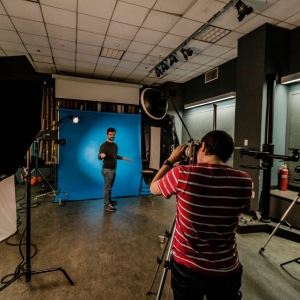 Interior of a photo studio. a person in the foreground is looking through a camera on a tripod at another person standing further back in the room against a blue seamless backdrop. 
