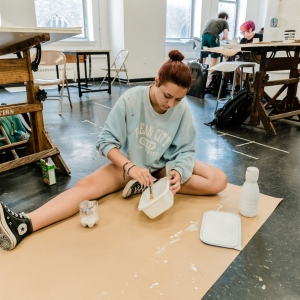 a person sits on the floor of a studio on top of sheet of paper and mixes a material in a white bowl. 