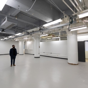 interior of new studio construction in Anderson hall. Stark utilitarian space with high open ceiling with pipes, wires, and fluorescent lights. The walls are bright white, as are three support pillars. There is a black door at the far right. the based of the wall has a black edging. A person wearing an all-black outfit and black face mask stands below the lights. 