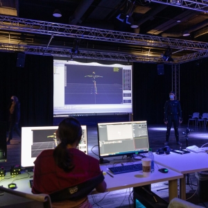 in the foreground, a person with a ponytail sits at a white desk facing two large computer monitors. beyond the desks is an open floor in a dusky room with supportive trusses across the length of the ceiling. a person in a body-mapping jumpsuit stands in the room. everything is bathed in a pale lavender light. in the background is a projector screen showing metrics of the body mapping