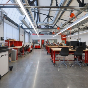 interior of UArts makerspace. an industrial-type space with a very high, slanted ceiling and windows for additional illumination. below the ceiling are ventilation pipes and long fluorescent lights. the floor is shiny and buffed, and throughout the space are wood workshop tables with red wheeled frames. 