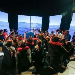 stage performance of I want a country. a mass of people in face masks sit in chairs around a group of people seated on the floor. the latter group is wearing red life jackets and many have their hands raised. all people are looking at hanging panels that surround the entire group, displaying an empty ocean in stark sunlight.