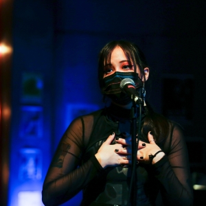 performance of we're gonna die. A person in a black sheer top with bright red eye makeup and wearing a black surgical mask clasps their hands over their chest. their face is close to a microphone. in the background, blurred light bulbs trail vertically along a red curtain, with the remainder of the background washed in blue light.