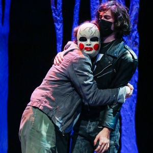 stage performance of hotspot. two people stand on stage awkwardly embracing against a background of blue velvet. the person on the right seems stiff, leaning slightly backwards in a black leather jacked and black surgical mask. the person on the left is clinging to the other, and has a creepy doll-like mask covering their face