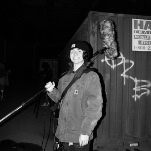 a black and white image of a person holding a boom mike on their shoulder and smiling at the viewer against a background of a metal shipping container sprayed with tags. 