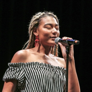 a person with bleached braids wearing a striped top and large dangly red earrings sings into a microphone with eyes closed against a completely black background. 