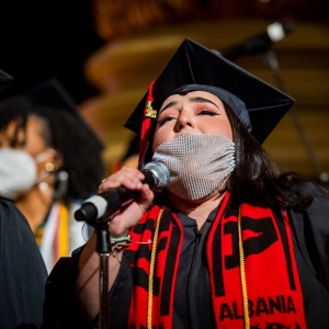 a person in a black graduation robe and cap sings into a mic through a sheer, gem-studded mask. they are wearing a red graduation stole that reads "albania" 