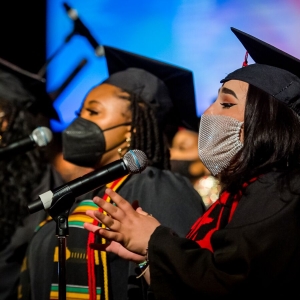 three people in black graduation robes and caps are seen in profile singing into microphones while wearing masks. the person in the middle has a kente-style graduation stole. the person in the foreground (at the right) has a sheer diamond studded mask and has their hands raised. 