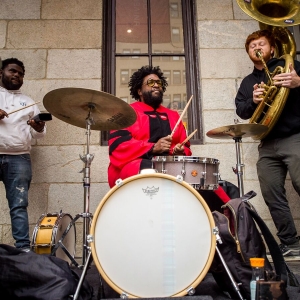 At the center of the image, Questlove is seated behind a drum kit wearing a crimson doctoral robe. Directly to the right, a student in a black shirt plays a tuba, as a student to the left in a white hoodie plays the cowbell. Image perspective from approximately the top of the kick drum. 