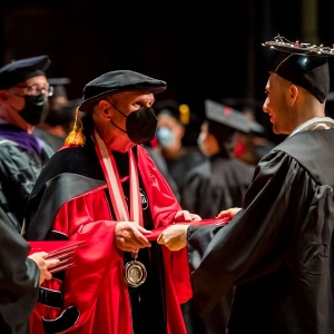A person on the left, seen from the front, is wearing a red doctoral robe, low-hanging gold medallion and a black face mask is handing a red leathery diploma to a person, seen from the back, in black graduation grab. The receiving person has a tangle of white lights on their graduation cap. To the left, a person holds a stack of diplomas. 