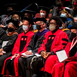 A crowd of seated people in face masks and doctoral robes look out at proceedings beyond the image. In the front row of people, three are wearing crimson robes, while the rest wear black ones. Many are holding booklets or binders with papers in their hands. 