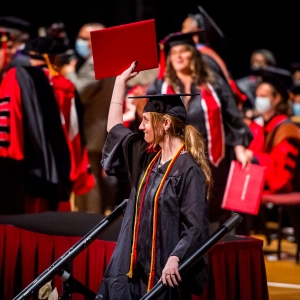 A person in a graduation robe with red and yellow stoles steps down from a stage, bites their lip, and holds a red leather-bound diploma aloft. In the background, people in graduation regalia mingle. 