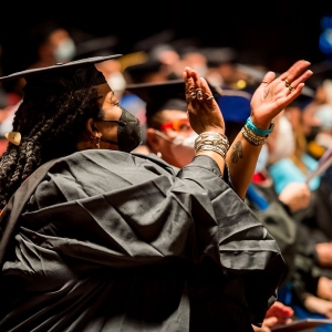a person in a black graduation robe, seen from the side, claps enthusiastically while looking off into the distance. The person has many bangles and rings on their hands and is wearing a black face mask. There are many seated people in varying graduation garb seen out of focus in the background. 