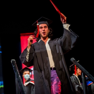 A person in an unzipped graduation robe shouts for joy and holds a diploma of their head while stepping down from a stage. A crimson UArts banner and an azure projection are seen out of focus in the background. 