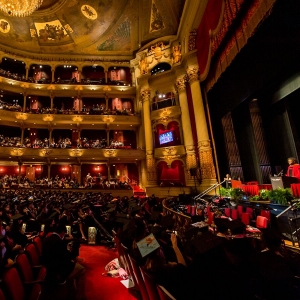 A view across a large, 4-story gilded auditorium that is brimming with people during a graduation ceremony. The ceiling is adorned with gilded paintings and a glowing chandelier, red velvety textures line the seating, floors and walls. A person in a crimson doctoral robe stands on a foliage-adorned stage at the right of the image and addresses the crowd. 
