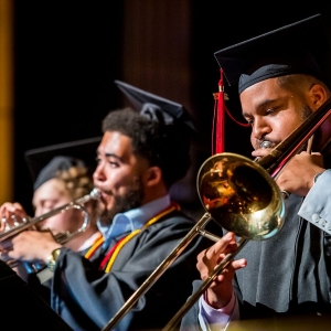 A row of students dressed in black cap and gown graduation regalia over pale blue dress shirts perform on brass instruments. The closest student on the right plays a trombone, the student in the center plays a silver trumpet.