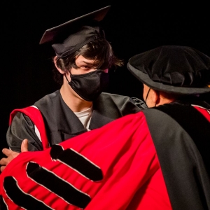 a student in a black graduation cap and gown and wearing a black face mask faces a person in a crimson doctoral robe, seen from the back and over the left shoulder, who is resting their hand on the students arm in a congratulatory gesture. 