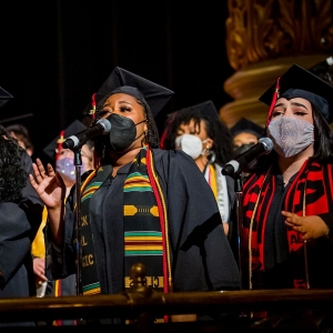 Three people wearing black cap and gown graduation regalia sing into their own microphones on stands. The image is shot from below hip level, looking up at their faces. The person on the left has a red graduation stole labeled class of 2021. the person in the middle has a black face mask and a kente-style stole. The person on the right has a sheer, gem-studded face mask and a wide red stole. 