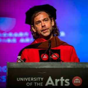 Aaron Dessner, seen squarely front the front, is dressed in a crimson doctoral robe emblazoned with UArts logos on the chest, and tilts his head and speaks into microphones at a podium labeled with a University of the Arts logotype.