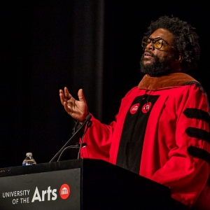 Questlove, seen angled from the right, is dressed in a crimson doctoral robe emblazoned with UArts logos on the chest, raises his right hand and speaks out over two microphones at a podium labeled with a University of the Arts logotype.